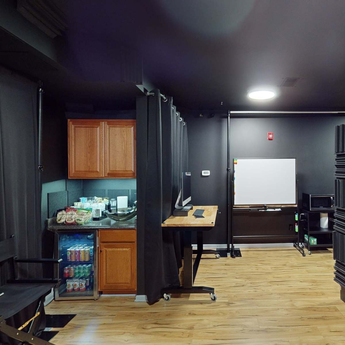 A room featuring a kitchenette with wooden cabinets, a whiteboard, and soundproof wall panels, all illuminated by a ceiling light.