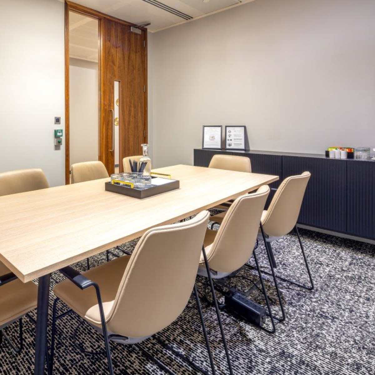 A modern meeting room features a long wooden table surrounded by eight beige chairs, with a coat rack and a textured rug on the floor.
