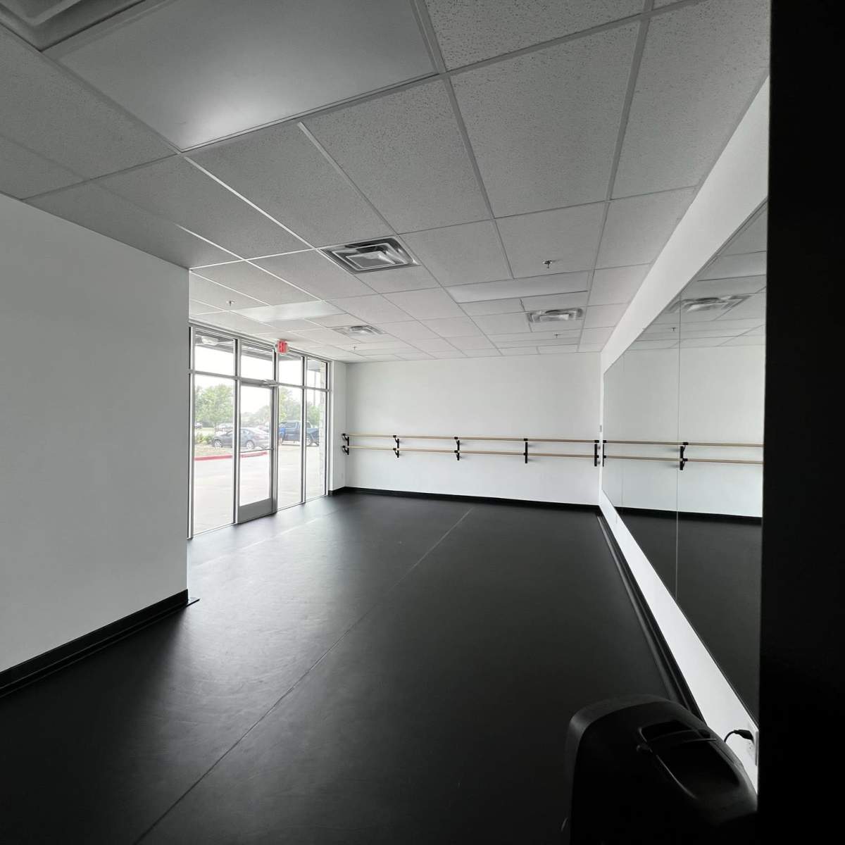 An empty dance studio with black flooring, white walls, and a large mirror along one side.