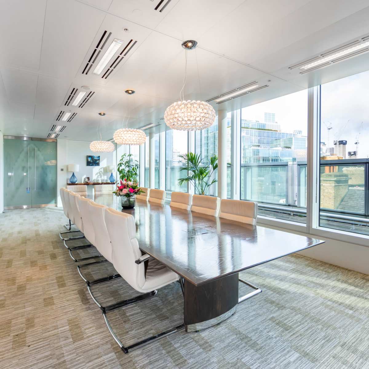 A modern conference room features a large wooden table surrounded by white leather chairs, with floor-to-ceiling windows and contemporary lighting.