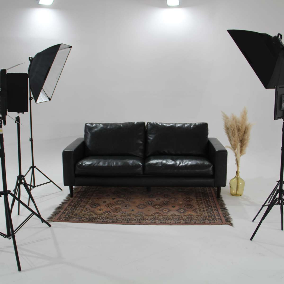 A black leather sofa is positioned on a patterned rug in a well-lit studio environment, flanked by two softbox lights and a decorative vase with dried plants.
