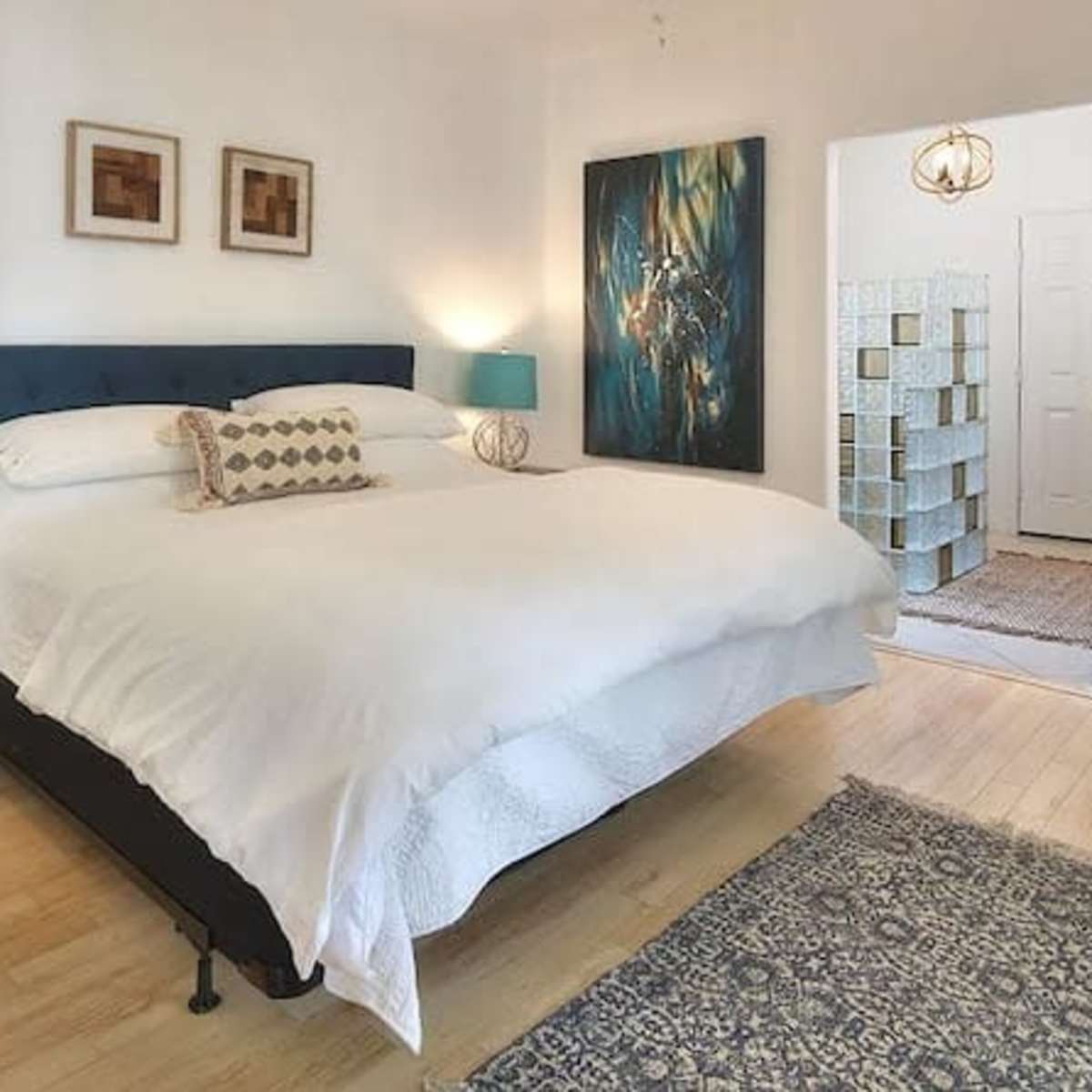 The bright bedroom features a large bed with white linens, two bedside lamps, artwork on the walls, and a textured rug on a wooden floor.