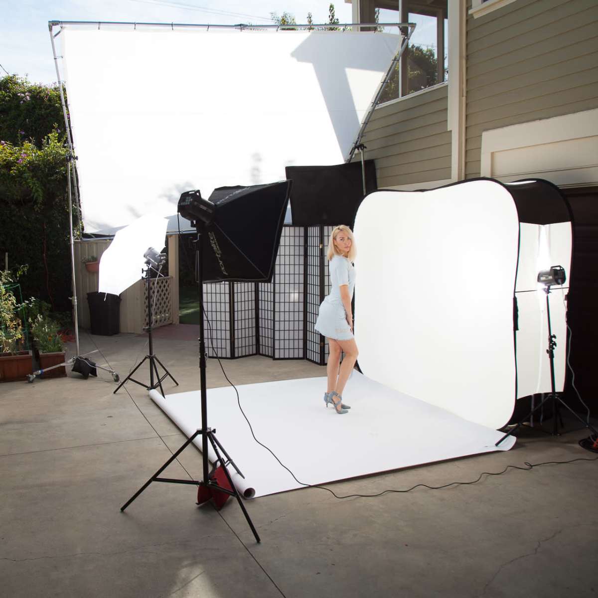 A photography studio setup is arranged outdoors, featuring a model posing in front of a white backdrop, with lighting equipment and plants in the background.