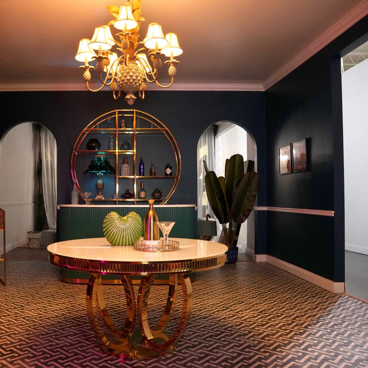 A stylish room featuring a round table with a gold base, a chandelier, and a bar area decorated with bottles and plants.