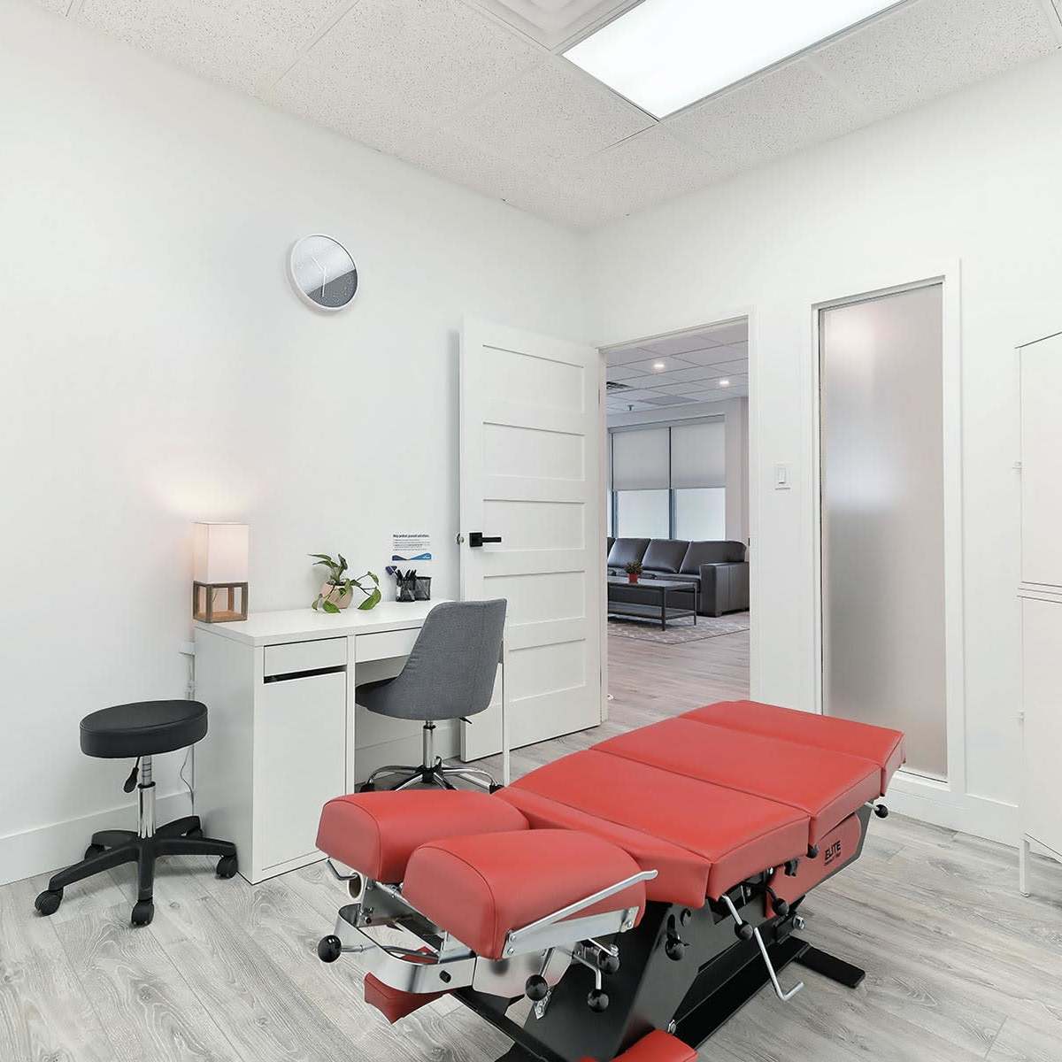 A clean examination room featuring a red treatment table, a small desk with a chair, a sink, and a storage cabinet, all set against white walls and a light wooden floor.
