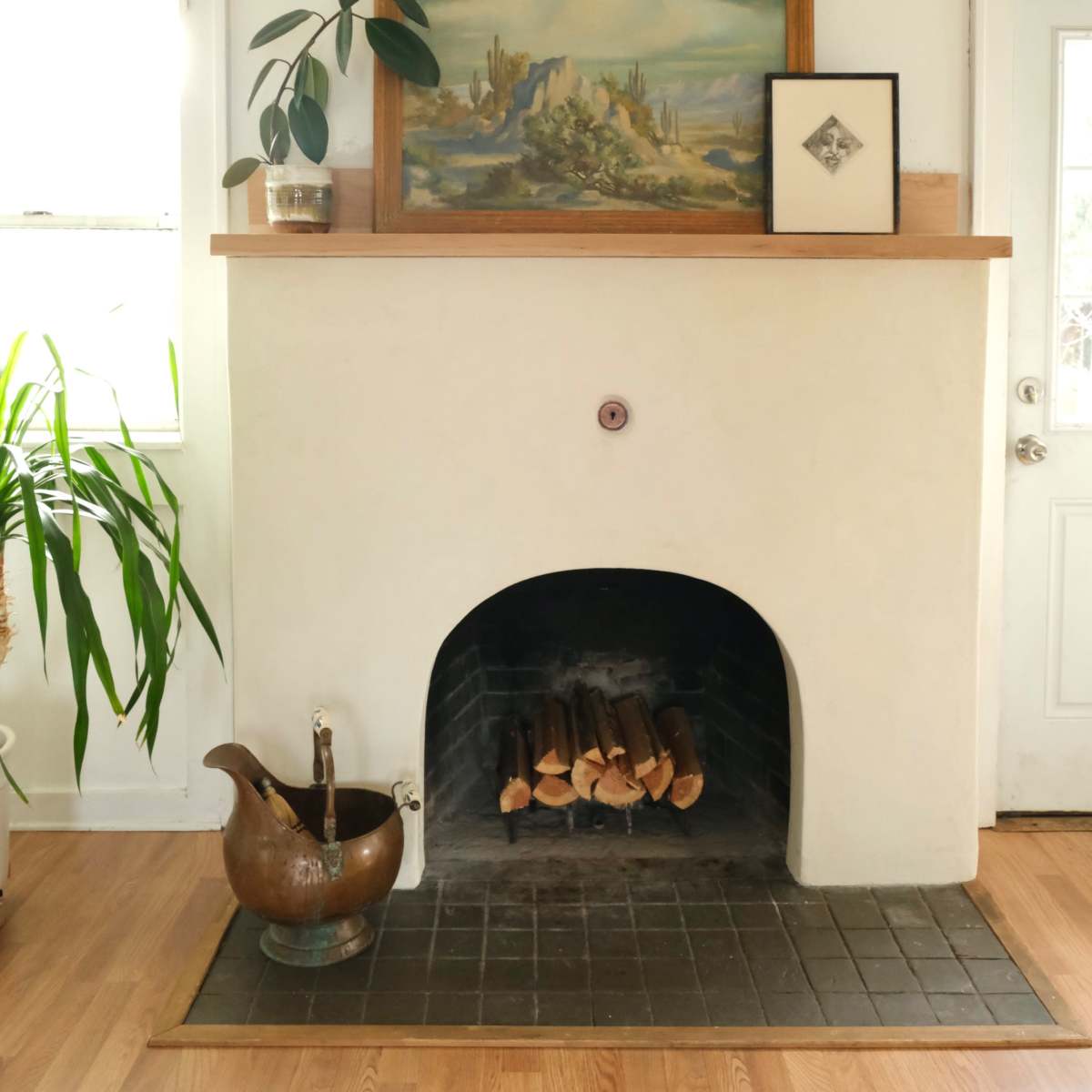 A white plaster fireplace with a wooden mantel holds a landscape painting, a framed artwork, and a potted plant, with logs stacked below and a decorative bowl beside it.