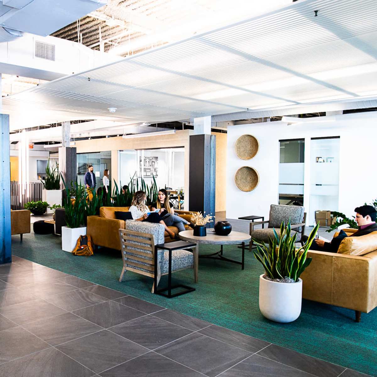 A spacious office lobby features comfortable seating areas with plants, large windows, and modern decor.