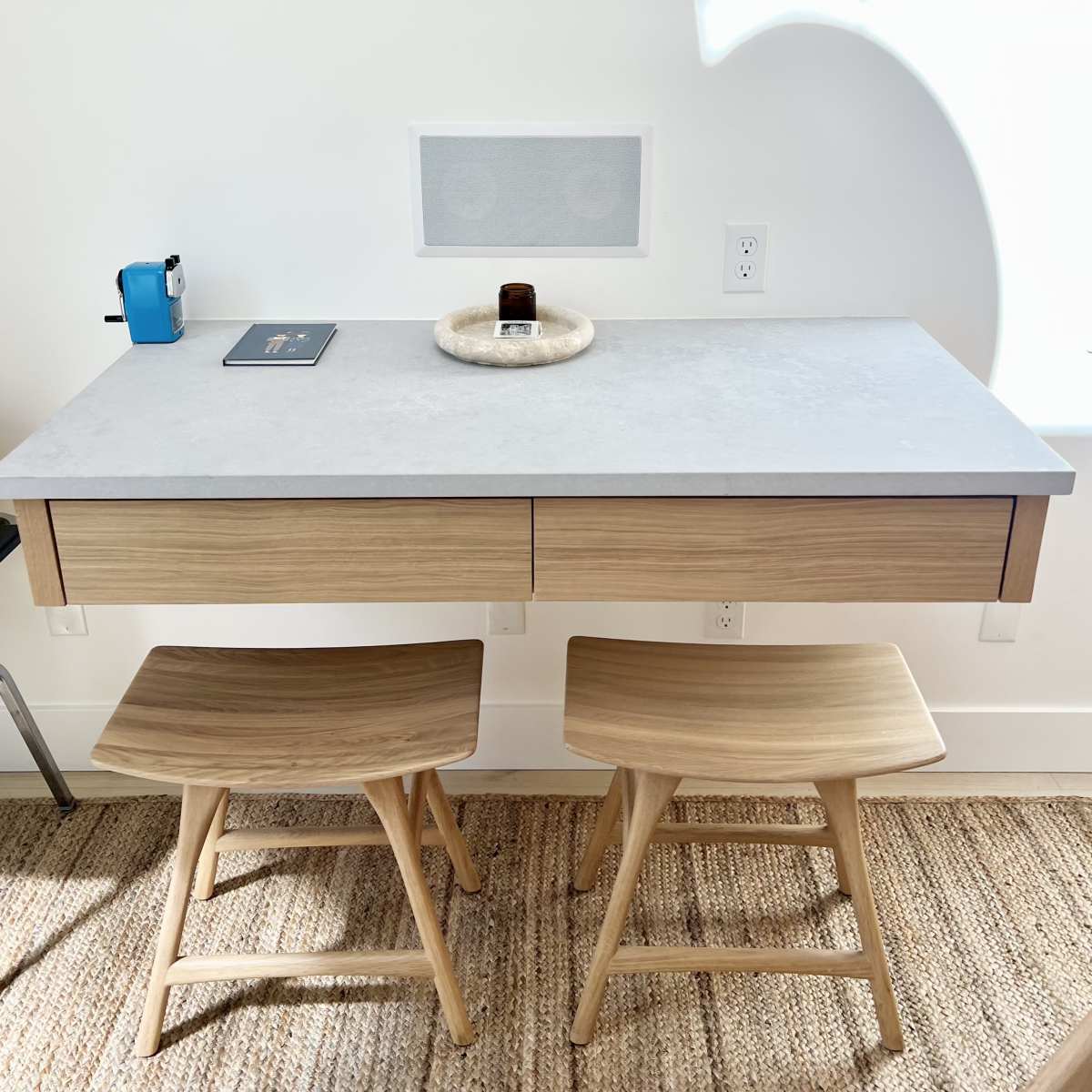 A wooden desk with two chairs and a small decorative dish sits against a white wall with a speaker and a window above it.