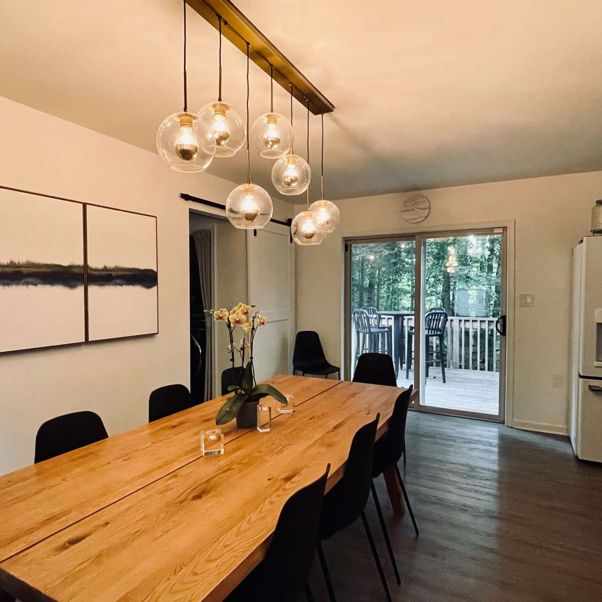 A dining area features a wooden table surrounded by black chairs, with pendant lights overhead and sliding doors leading to a deck in the background.