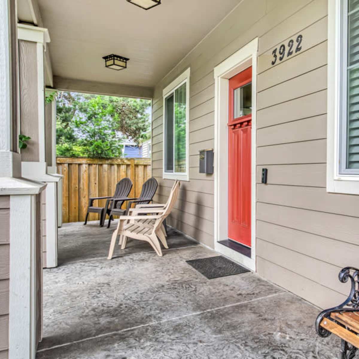 A covered porch with two wooden chairs and a bench, featuring a bright red front door and a fenced yard in the background.