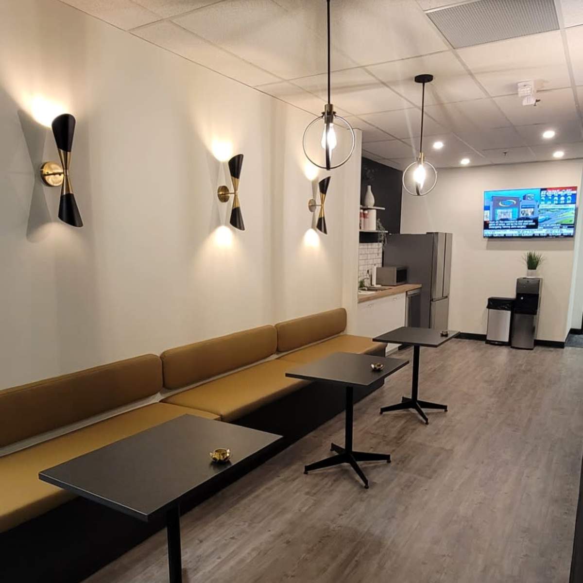 A modern office break room with a long bench, two coffee tables, pendant lights, and a television on the wall.