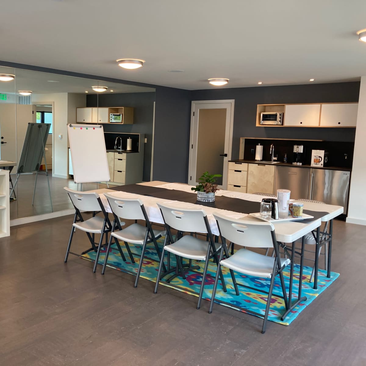 A modern meeting room with a long table set for several people, a large television on the wall, and a kitchenette area in the background.