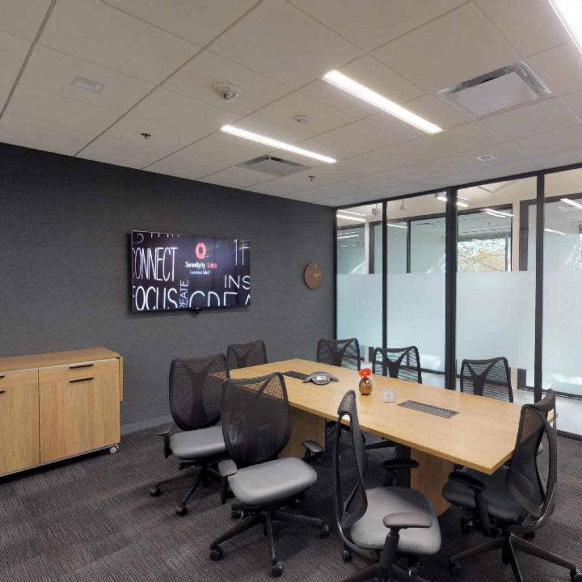 A modern conference room features a large wooden table surrounded by ergonomic chairs, with a wall-mounted screen and glass partitions.