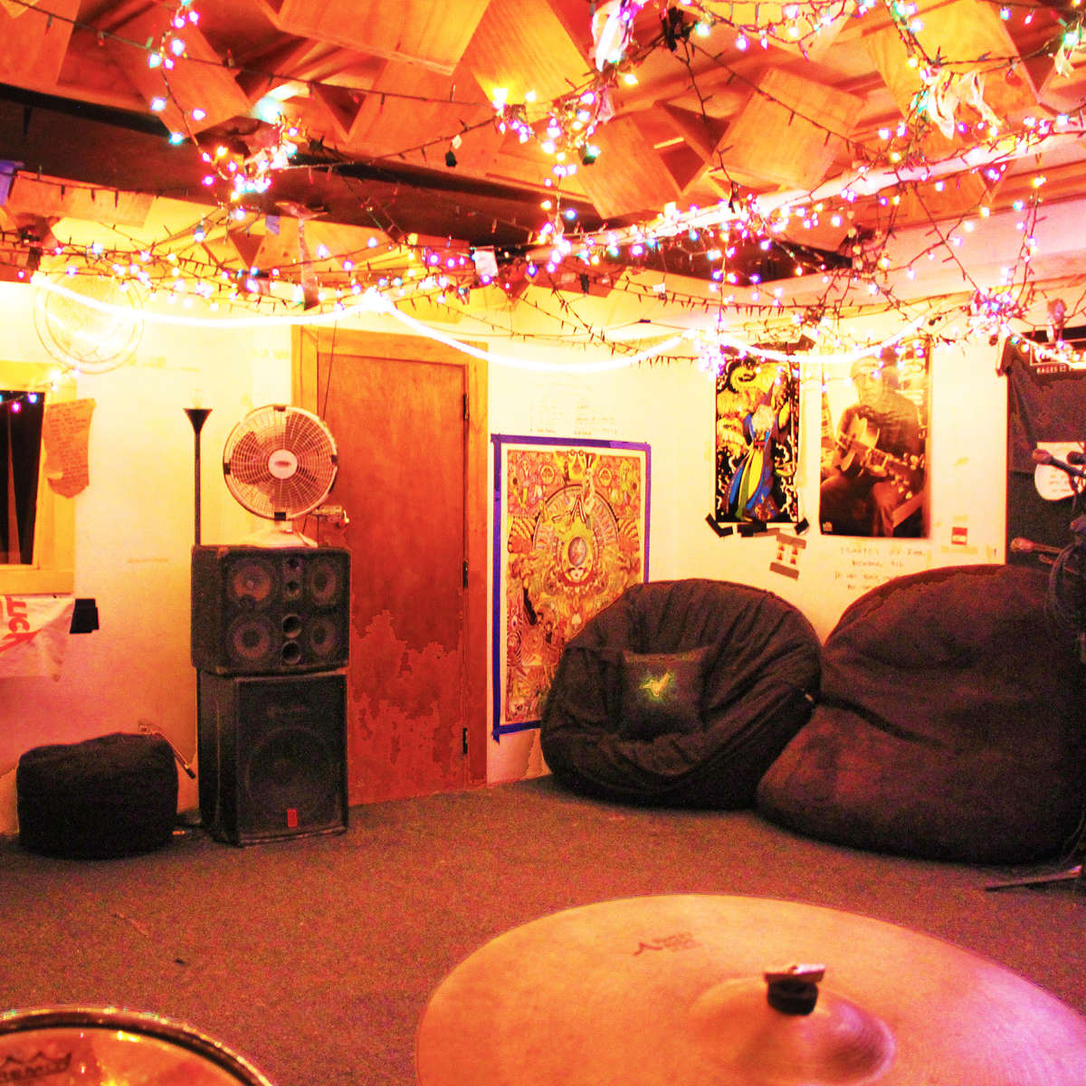 A music practice space with colorful string lights, a drum set, large bean bags, and a wall decorated with posters.