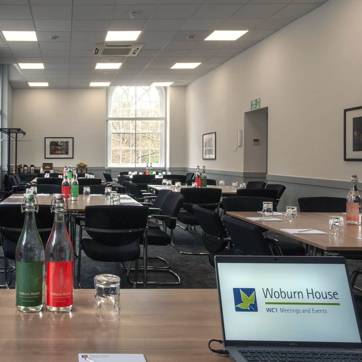 A conference room is set up with multiple tables and chairs, featuring refreshments on the side and a laptop displaying event information.