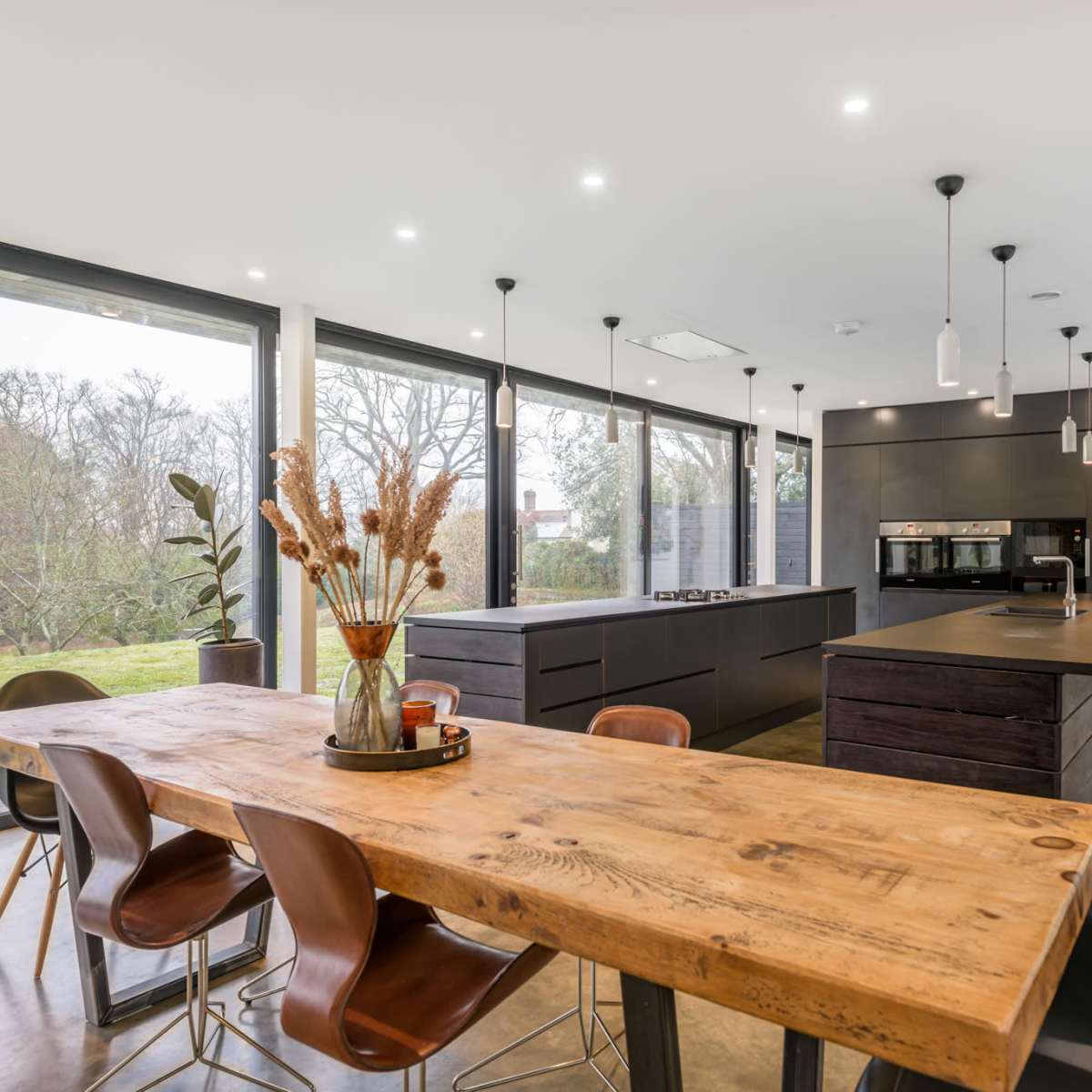 A modern kitchen features a large wooden dining table surrounded by chairs, sleek cabinetry, and large windows overlooking a green landscape.