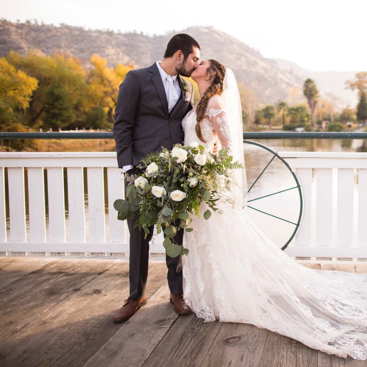 A bride and groom share a kiss on a wooden dock by a serene body of water, surrounded by autumn foliage.