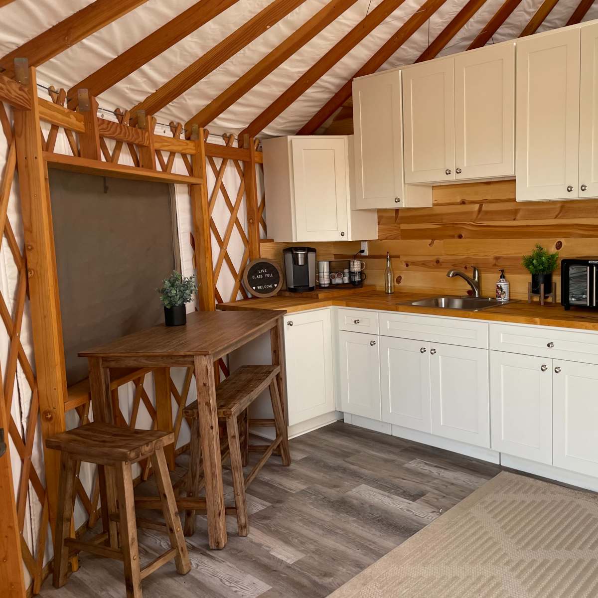 A compact kitchen area inside a wooden structure, featuring white cabinets, a countertop with a sink, a coffee maker, a microwave, and a refrigerator, along with a small table and stools.