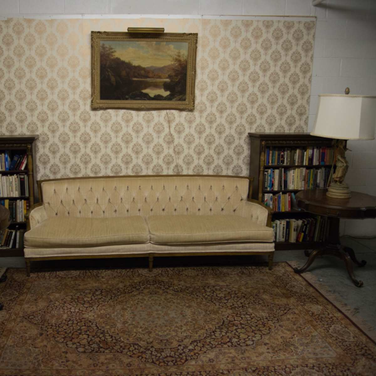 A vintage living room featuring a tufted beige sofa flanked by two decorative lamps on side tables, with a patterned wallpaper and a landscape painting above.