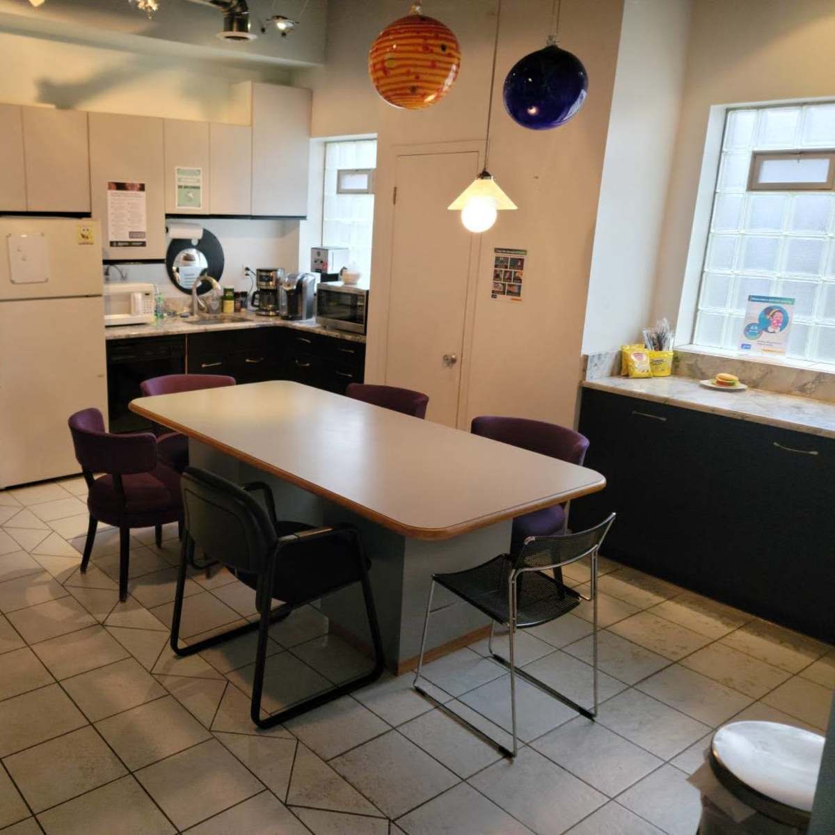 A kitchen with a large table surrounded by chairs, featuring a refrigerator, coffee maker, and colorful hanging lights.