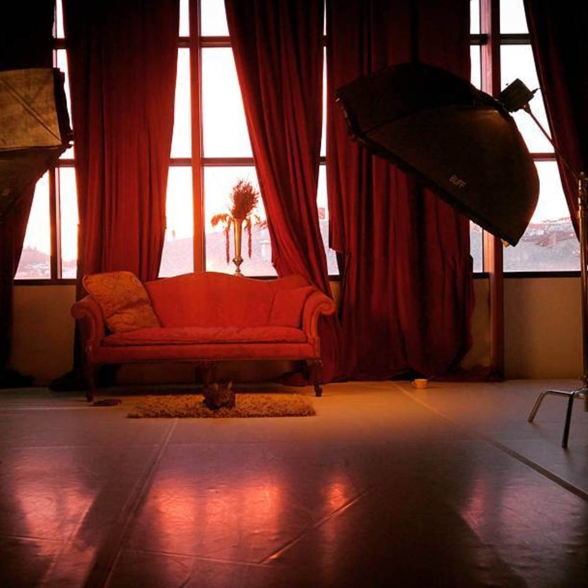 A pink couch is positioned in front of large windows, framed by red curtains, with two studio lights on either side.