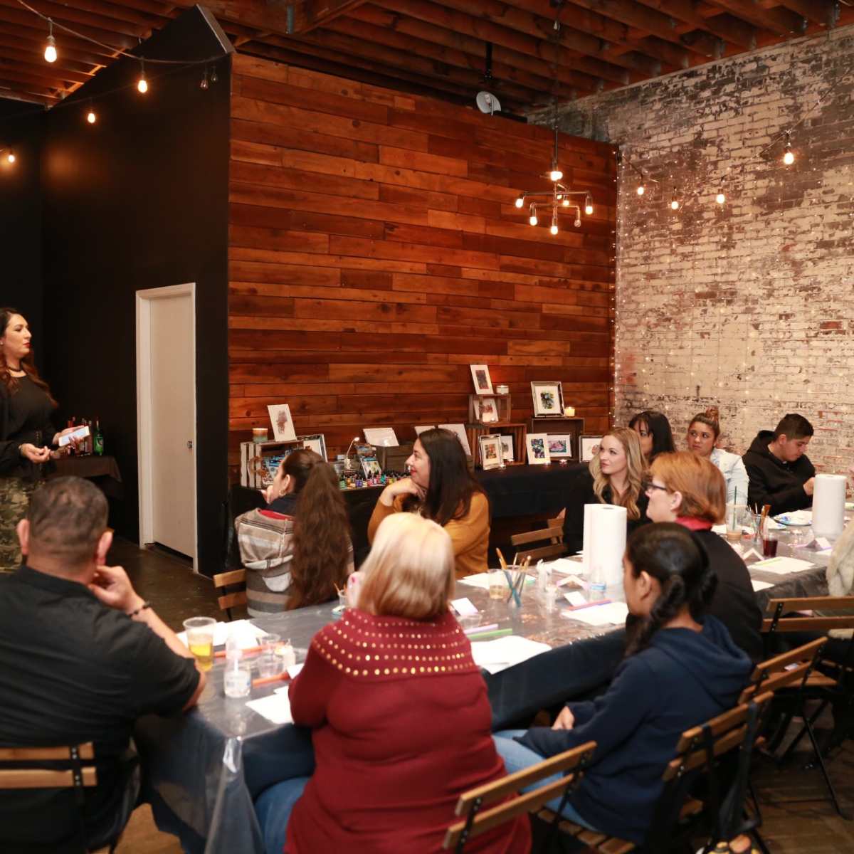 A speaker stands in front of a seated audience in a rustic venue, with wooden walls and string lights overhead.