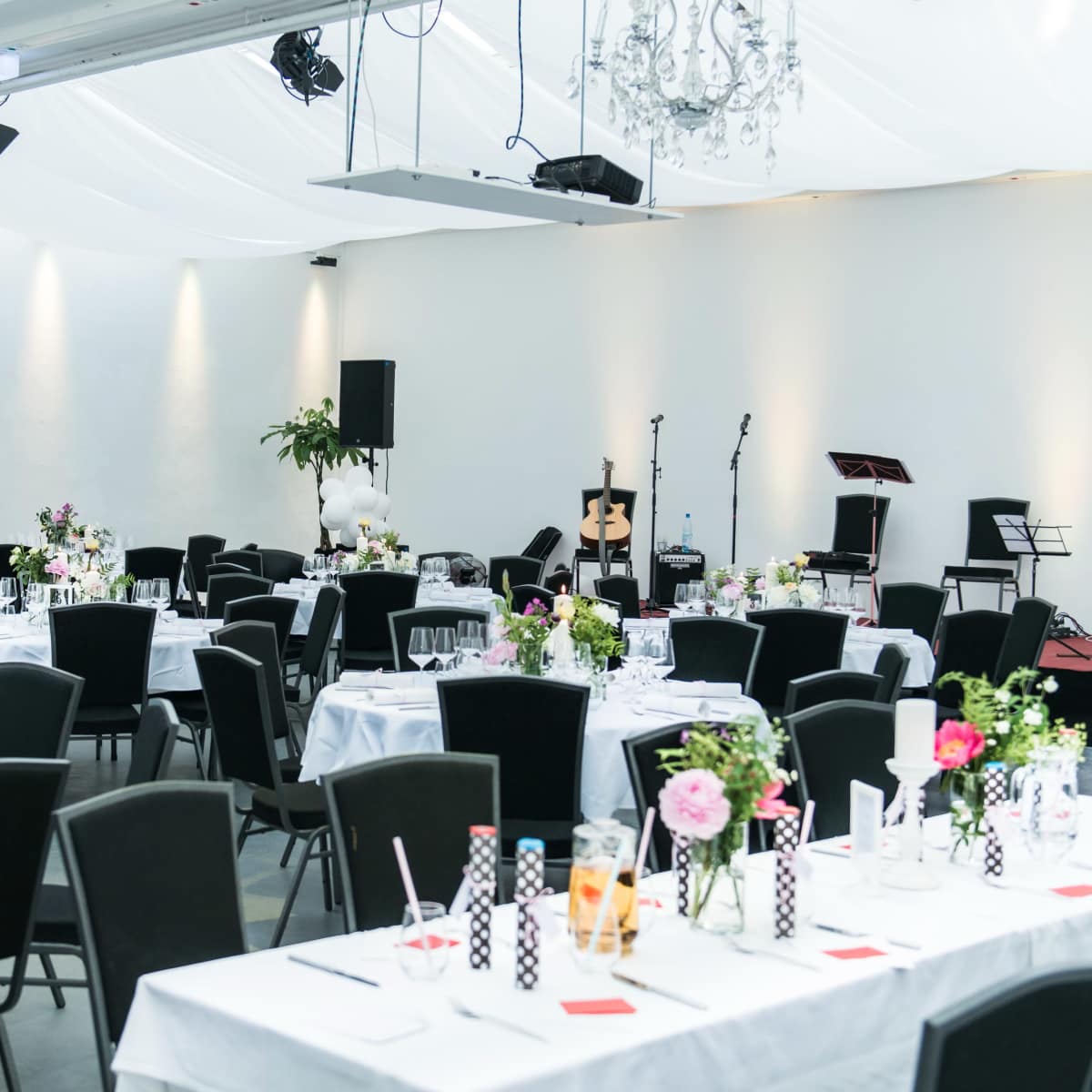 A banquet hall is set with elegantly arranged tables, each adorned with floral centerpieces and surrounded by black chairs, under soft lighting and decorative chandeliers.