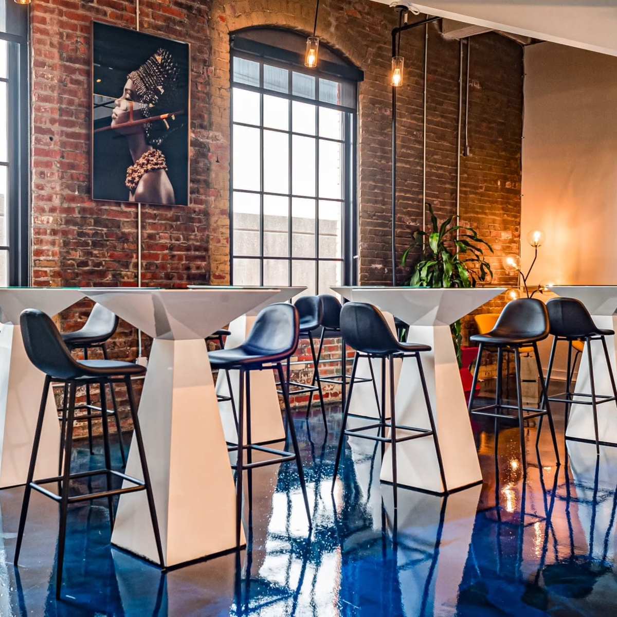 A modern interior with several white high tables and black bar stools against a backdrop of exposed brick walls and large windows.