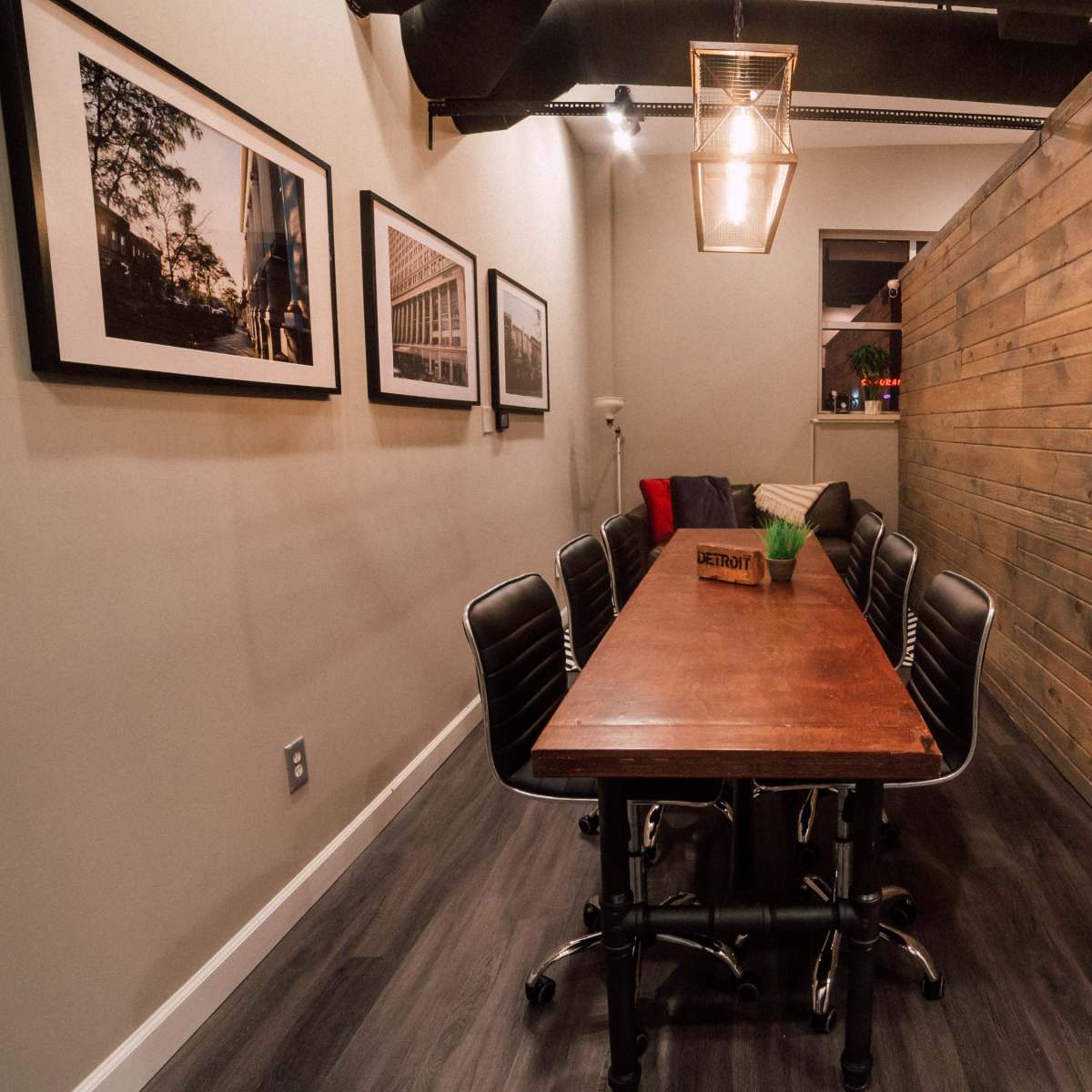 A long wooden table with black chairs is positioned in a narrow room featuring a wall made of wood planks and framed photographs on the adjacent wall.