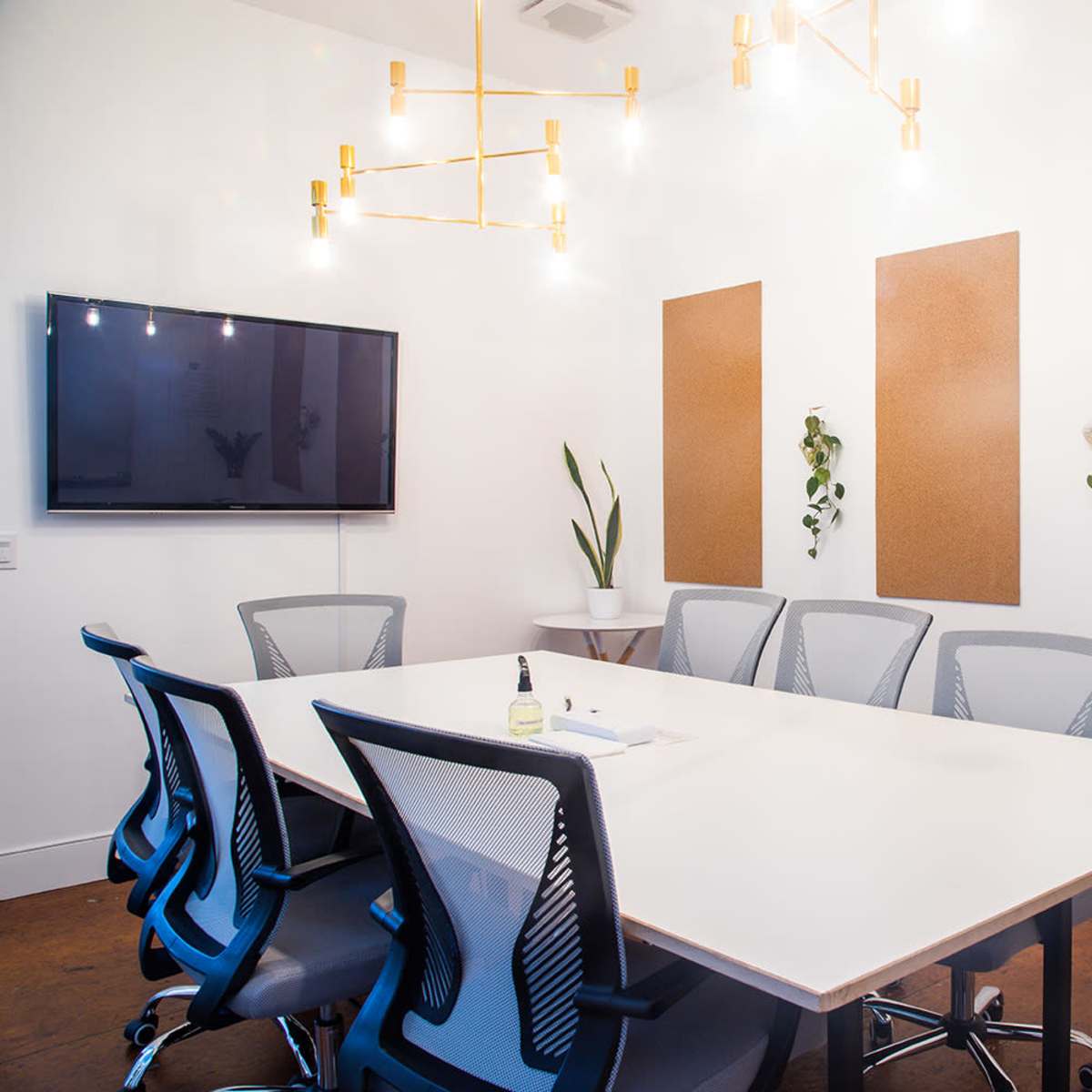 A modern conference room features a large table surrounded by ergonomic chairs, with a TV mounted on the wall and cork panels for notes.