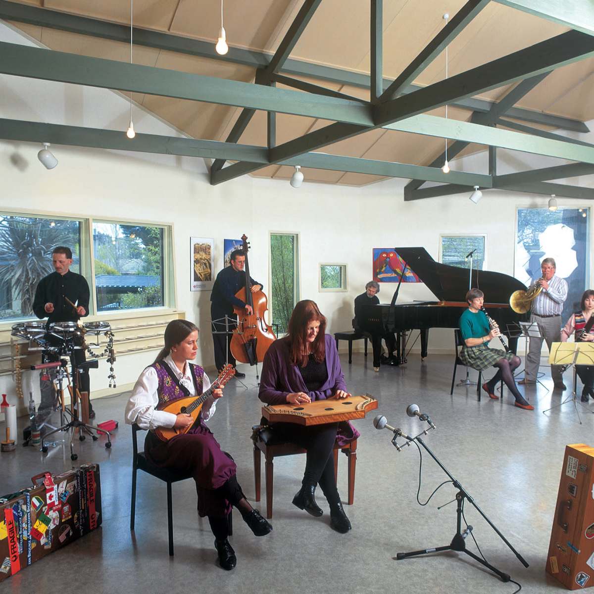 A group of musicians performs together in a brightly lit room with high ceilings and various instruments, including a piano, strings, and brass.