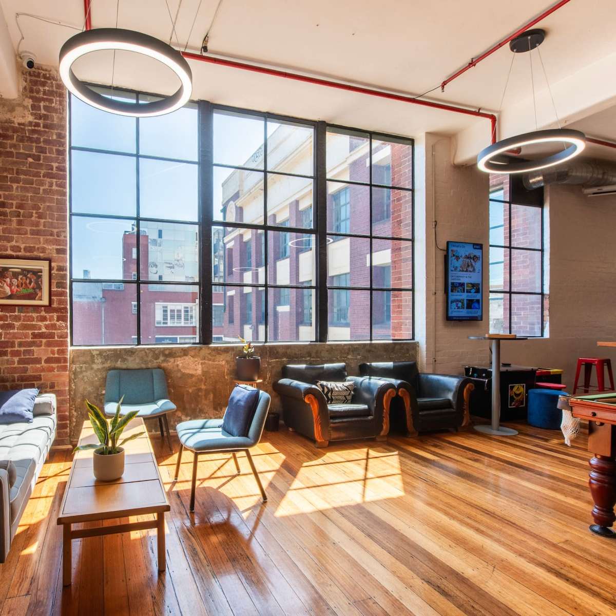 A modern communal living space with large windows, brick walls, and a mix of seating options, including a sofa and armchairs, illuminated by natural light.