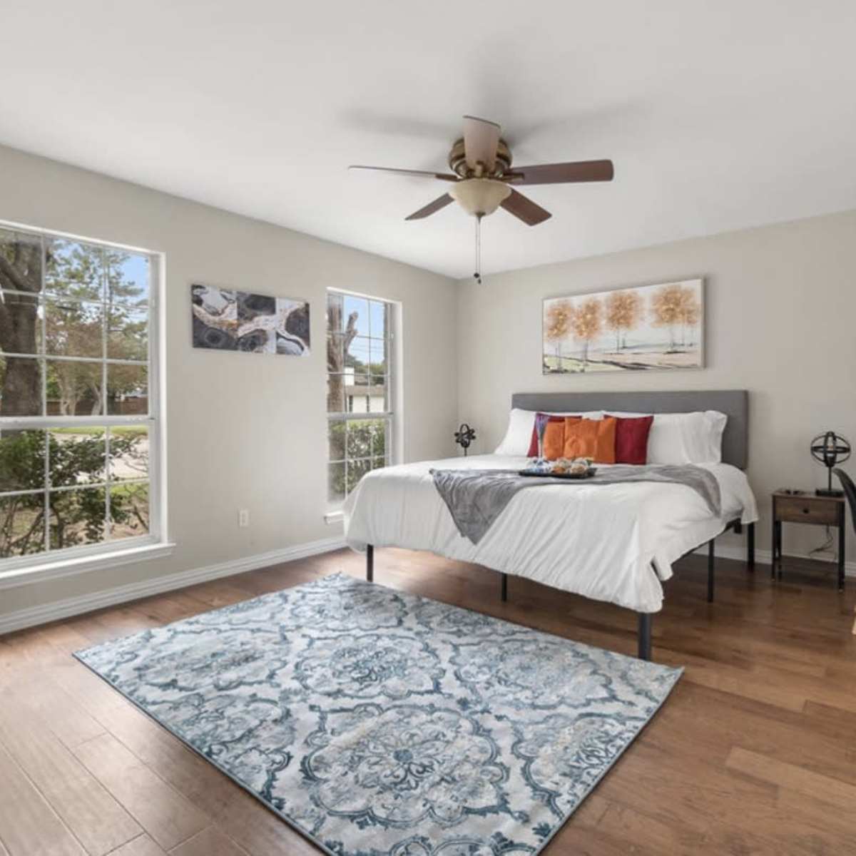 A spacious bedroom features a bed with decorative pillows, a ceiling fan, a large mirror, and a wooden desk next to a window.