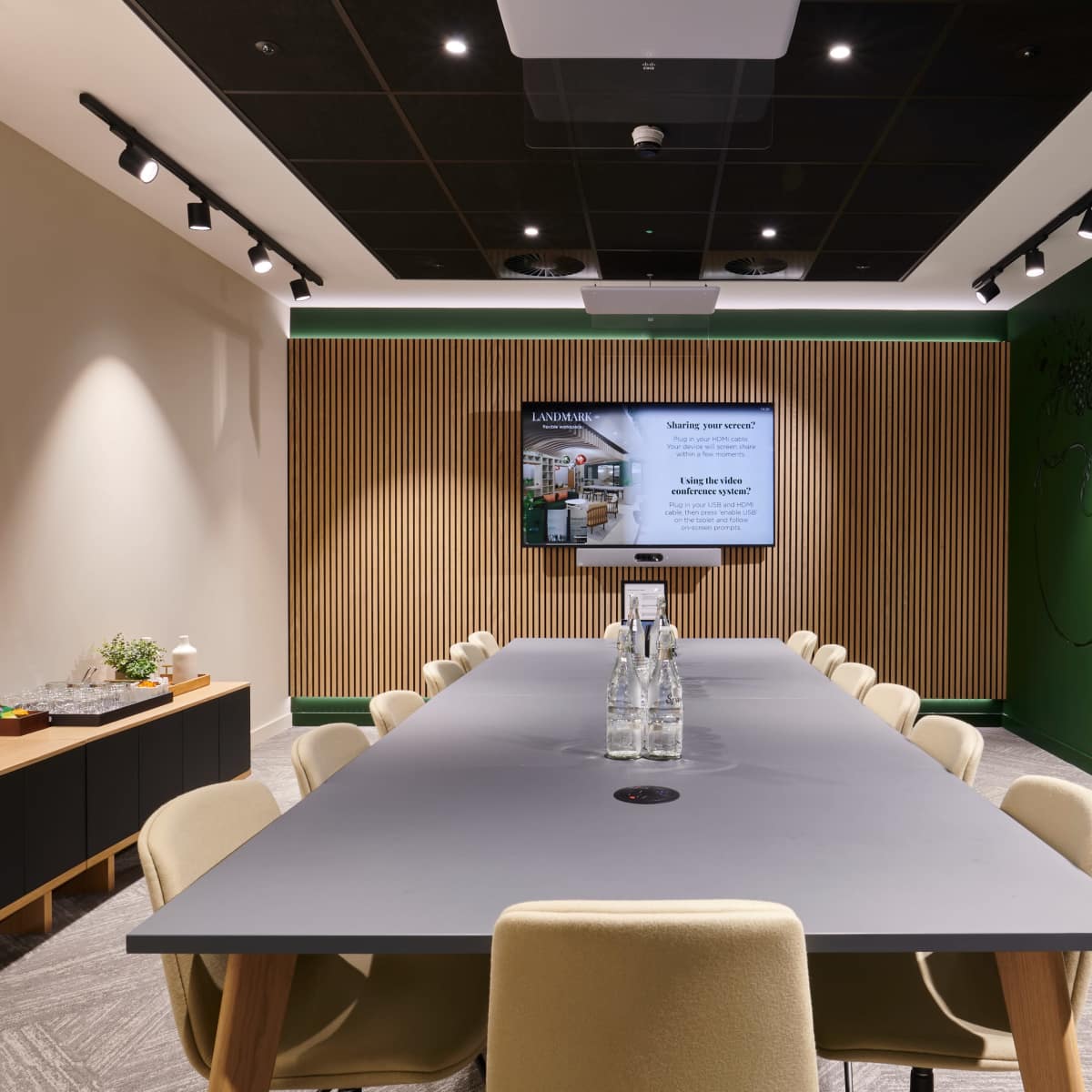 A long conference table with beige chairs is set in a modern meeting room featuring a TV and green accent wall.