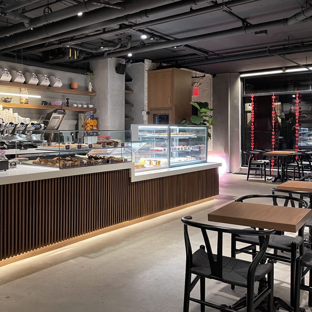 The interior of a modern café with a light wooden counter displaying pastries and a coffee bar, flanked by shelves of decorative items.