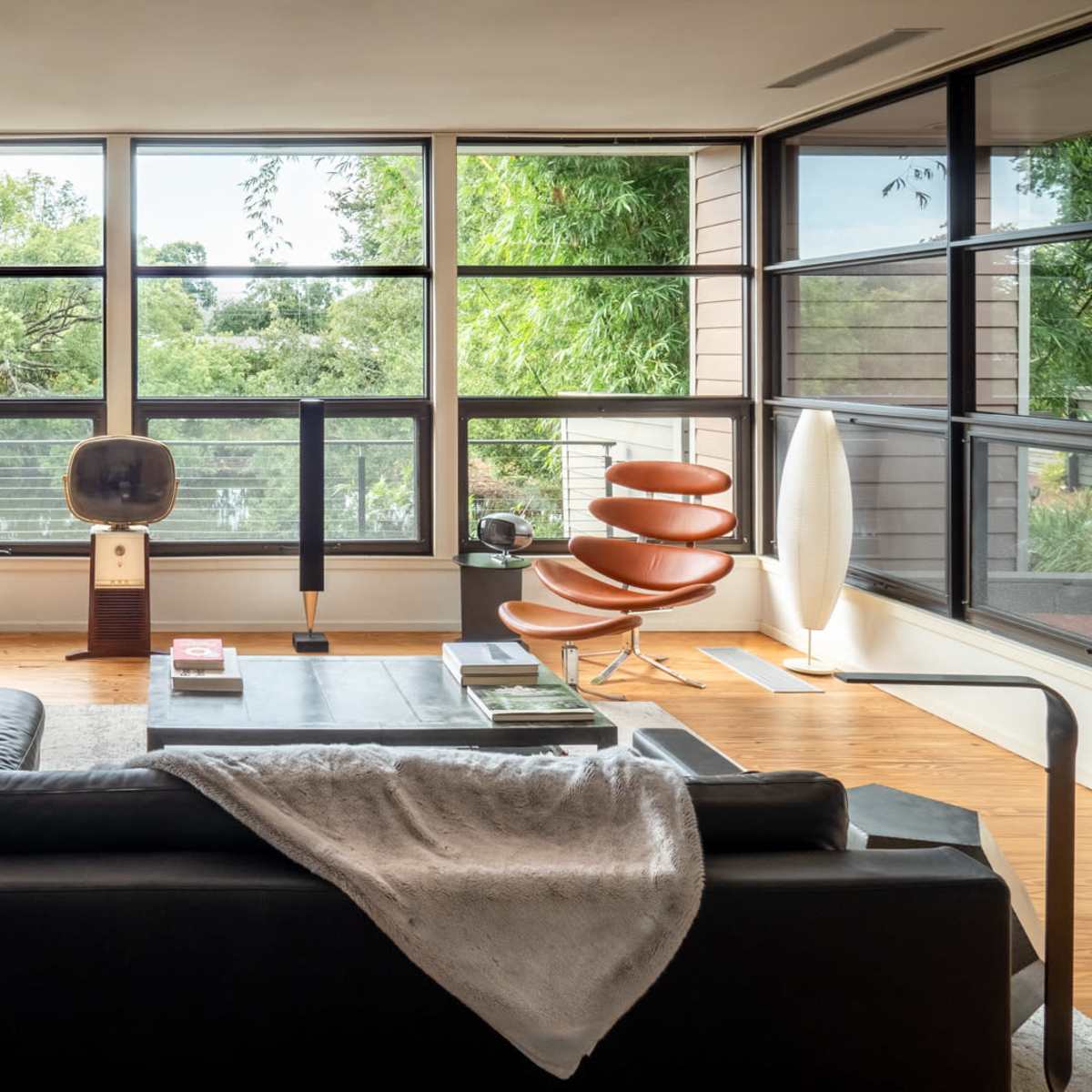 A modern living room with large windows, a black sectional sofa, and various mid-century modern furniture pieces.