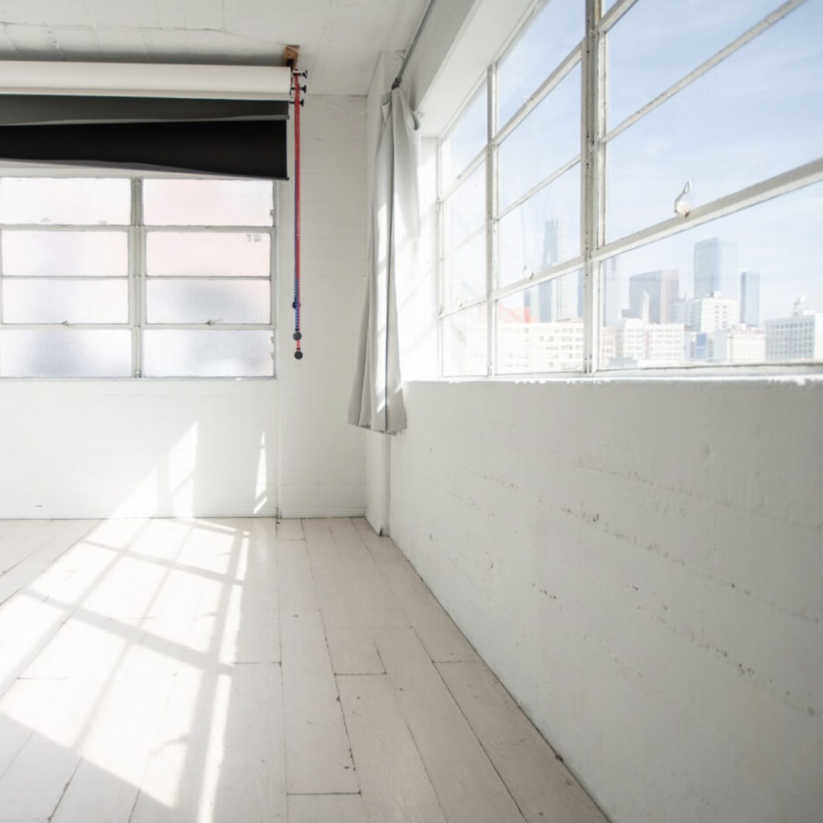 A bright, empty room with large windows and white walls, featuring a roll-up black curtain near the ceiling.