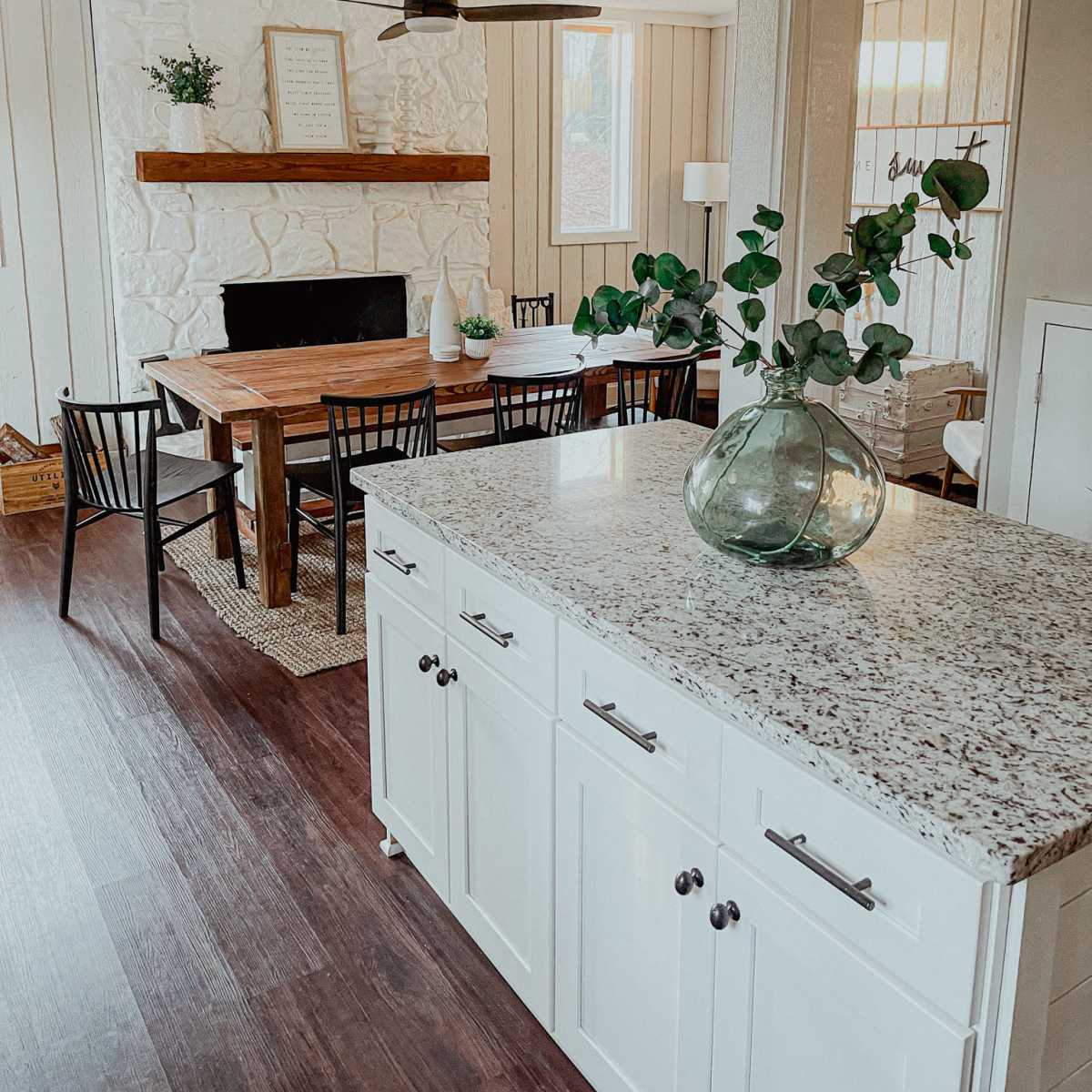 A modern dining area featuring a wooden table, black chairs, a stone fireplace with a wooden mantel, and a kitchen island with a granite countertop.