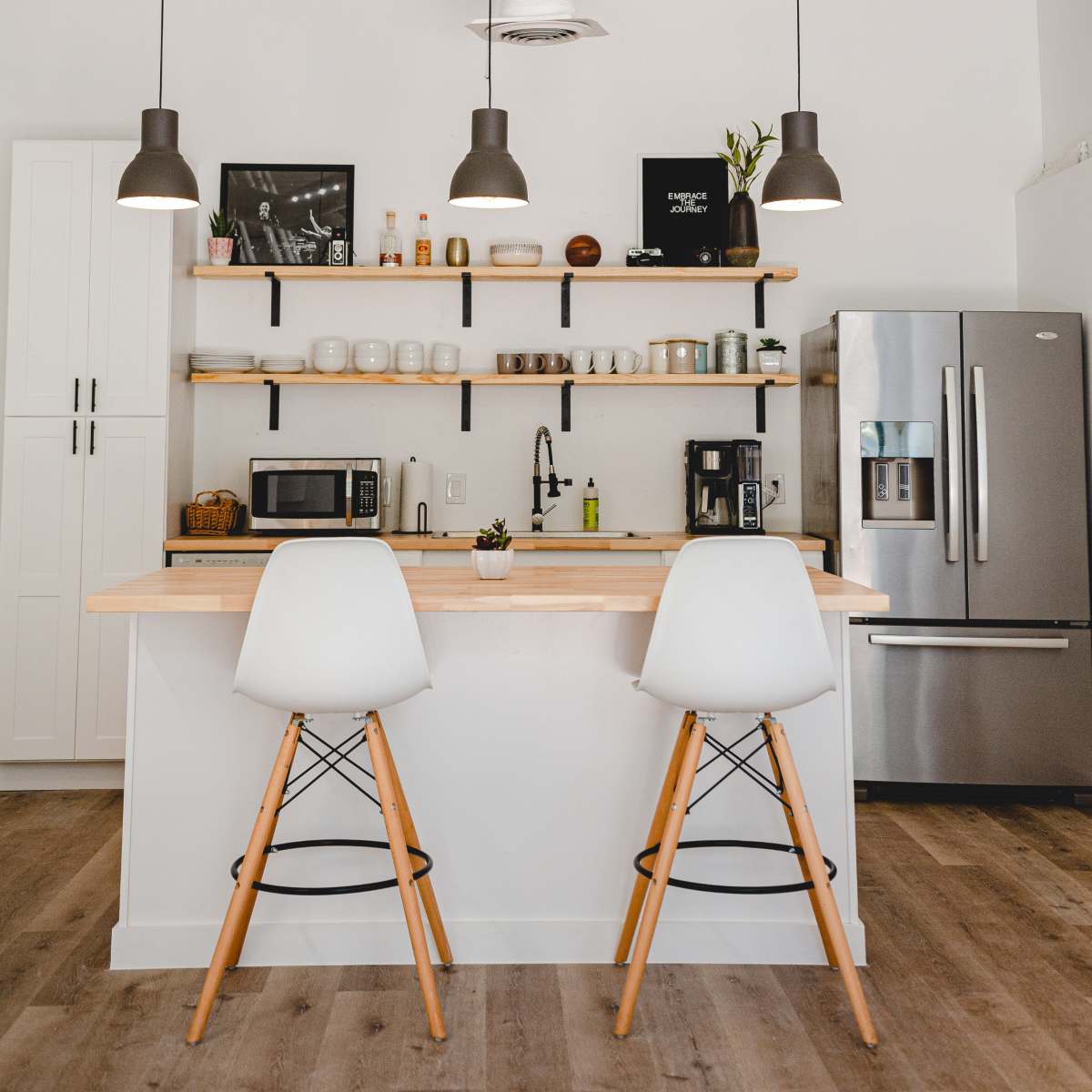 A modern kitchen features a bar with two white stools, a wooden countertop, open shelves displaying various kitchen items, and stainless steel appliances.