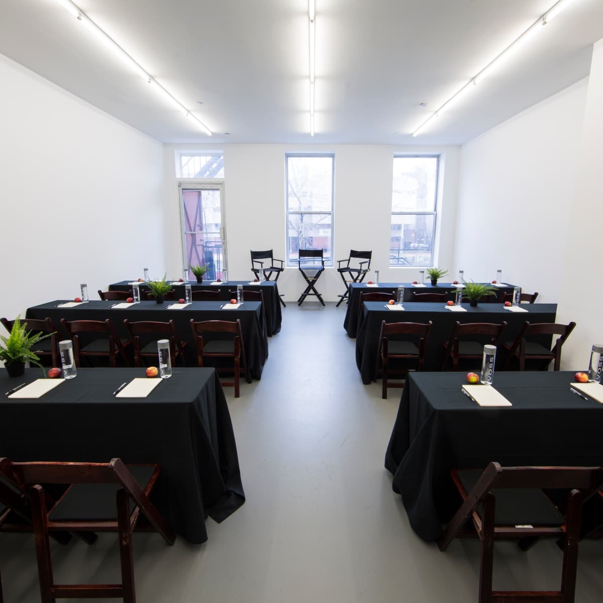 A spacious, empty room features two rows of tables with black tablecloths, each set with water bottles and writing materials, flanked by two chairs at the front.