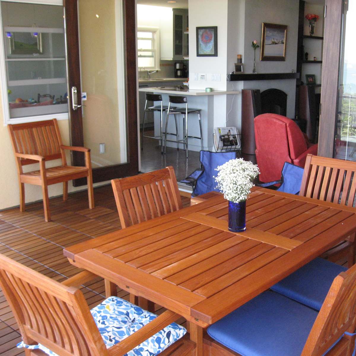 A wooden patio table with chairs is set up on a deck, overlooking an open space that leads to a kitchen and a view of the water.