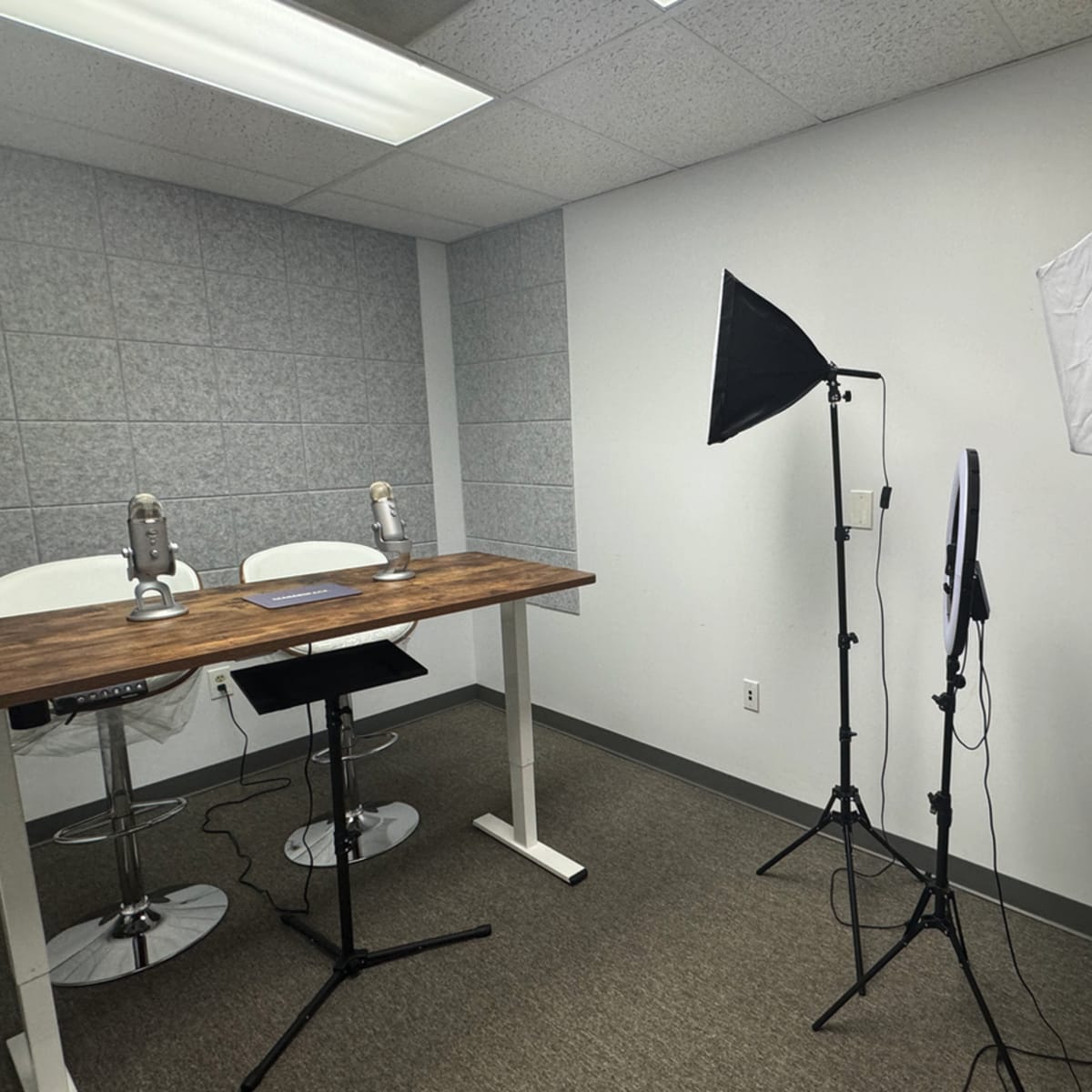 A minimalist recording studio features two microphones on a wooden table, flanked by lighting equipment.