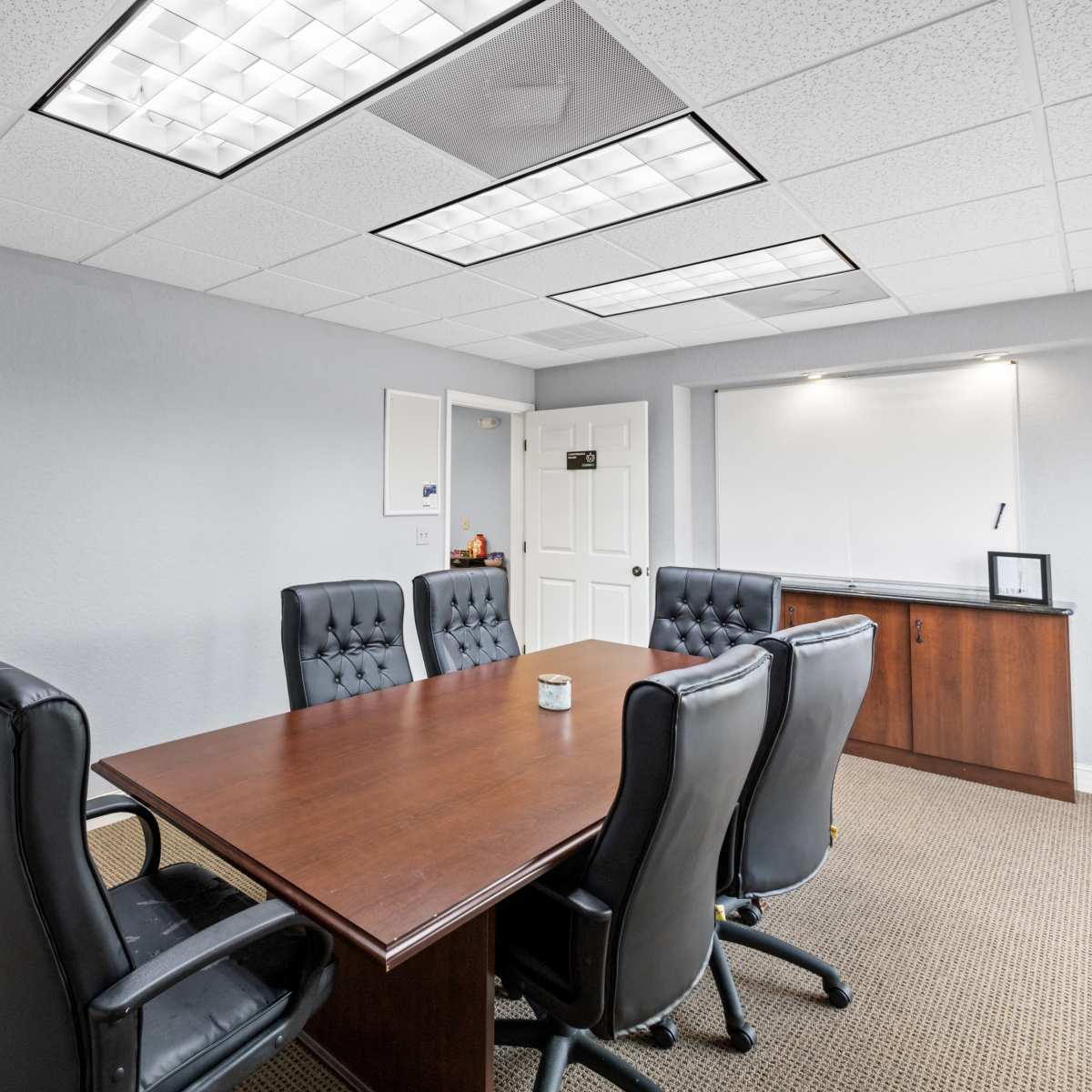 A conference room features a wooden table surrounded by five black leather chairs, with a whiteboard and a cabinet on the wall.