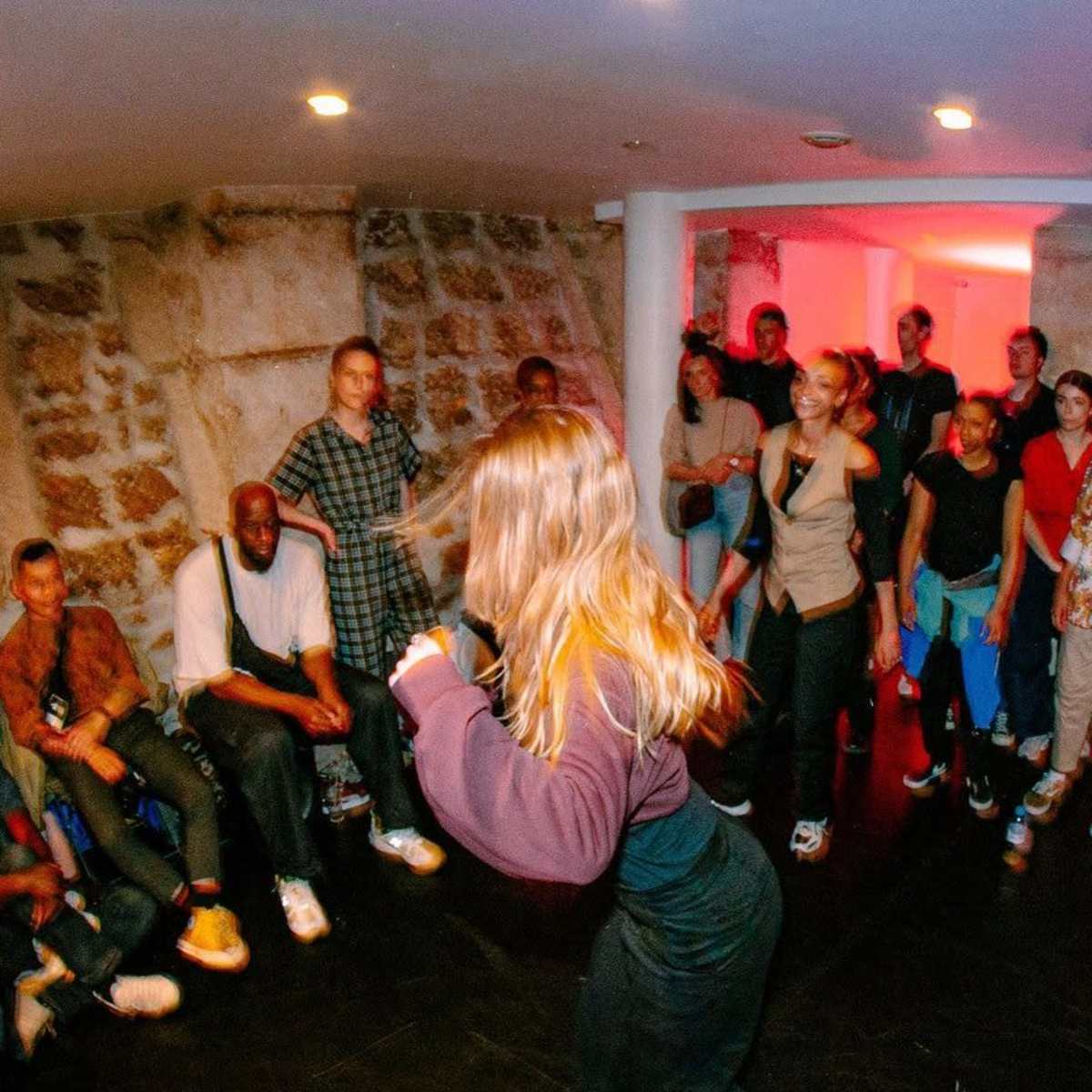 A group of people watches a dancer perform in a dimly lit basement venue with stone walls.
