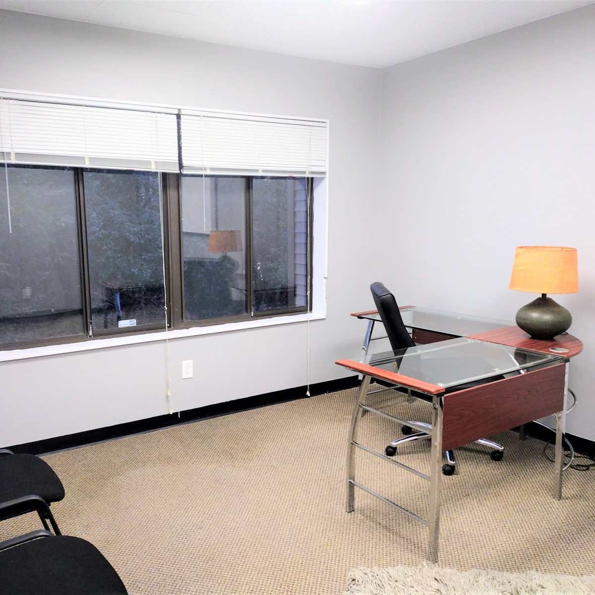 A simple office space with a glass-top desk, a lamp, and two black chairs against a neutral-colored wall.