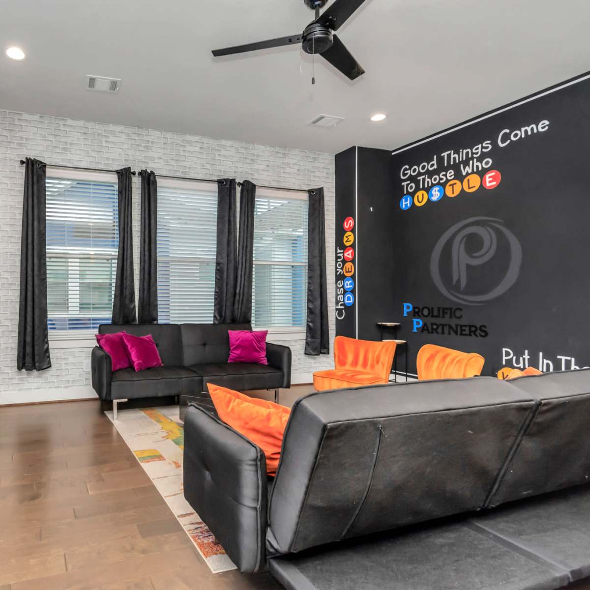 A modern living room with a black and white color scheme, featuring a large television, a dark sofa set, vibrant cushions, and a chalkboard wall with motivational phrases.