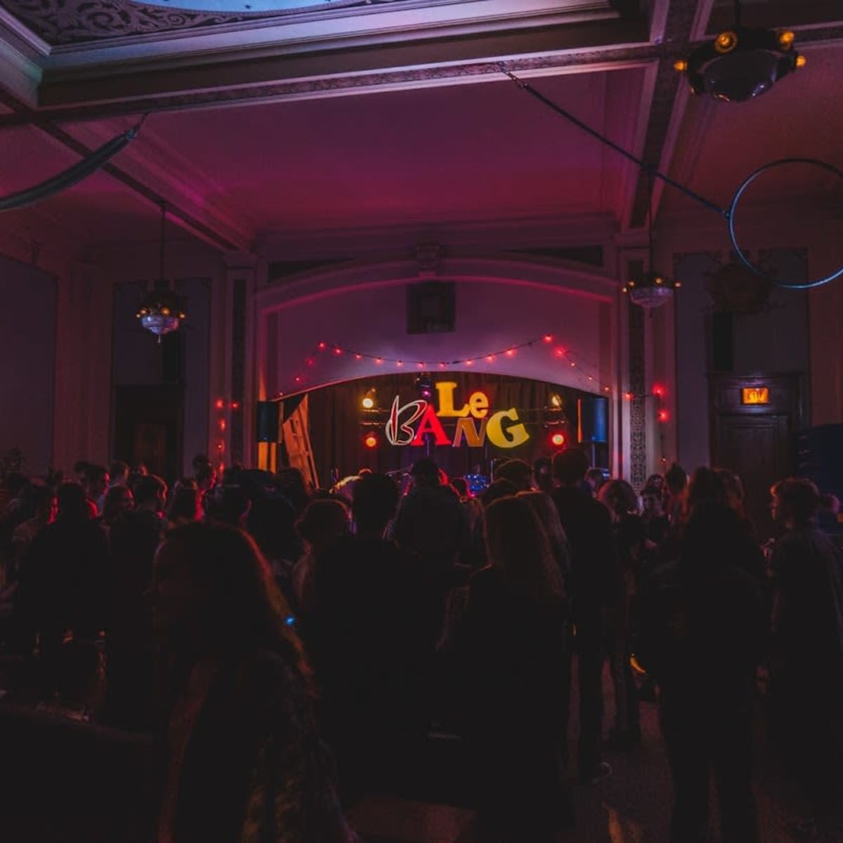 A crowd of people gathers in a dimly lit venue with colorful lights and a stage that displays the word "Le BANG."