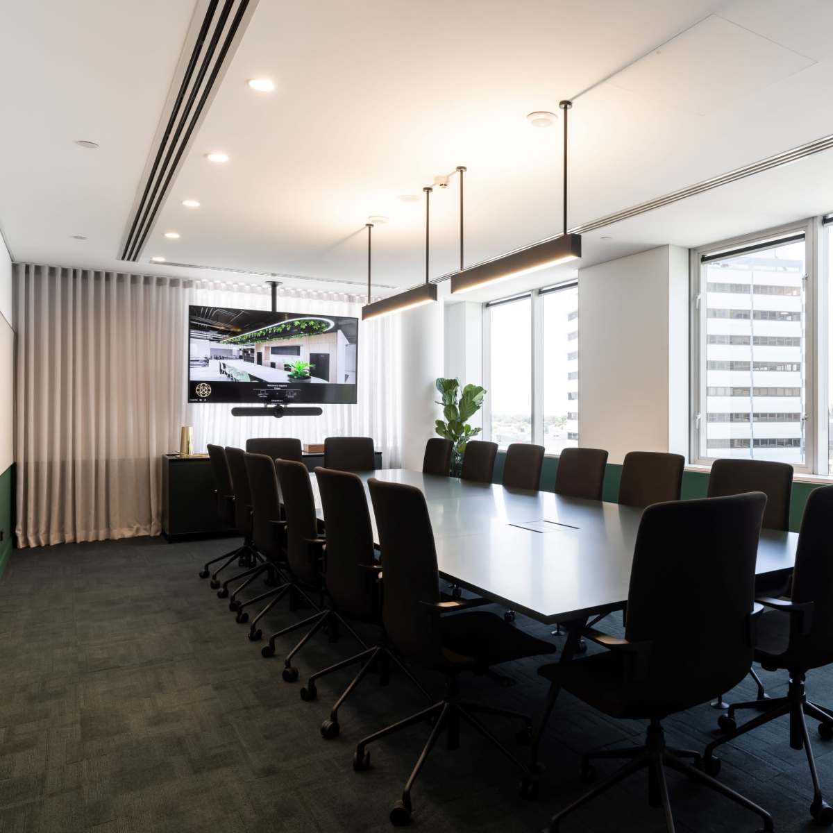 A modern conference room features a long table surrounded by black chairs, large windows with city views, and a wall-mounted screen.