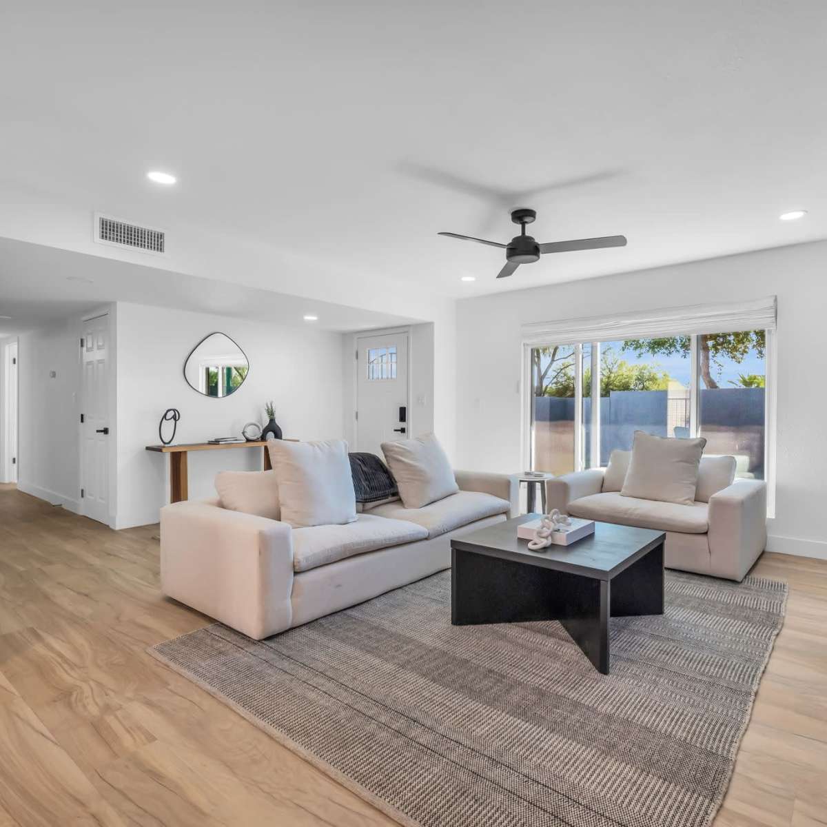 The room features a modern living area with a light-colored sofa set, a dark coffee table, and large windows that let in natural light.