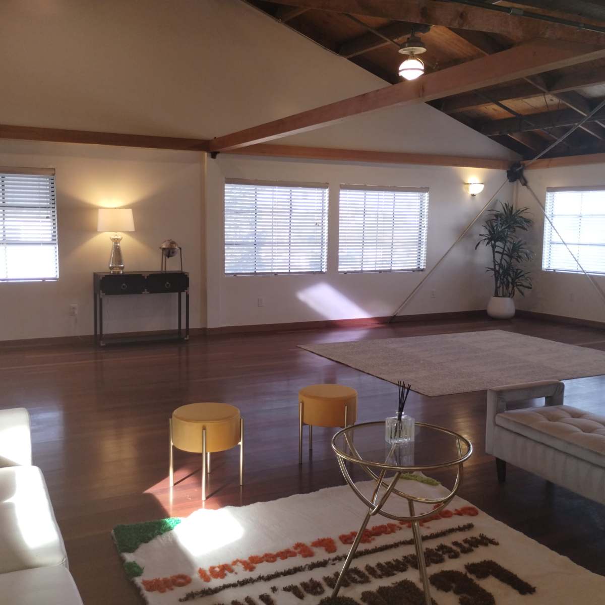 A spacious, well-lit room features large windows, light-colored walls, and wooden beams, furnished with a sofa, a coffee table, and small stools.